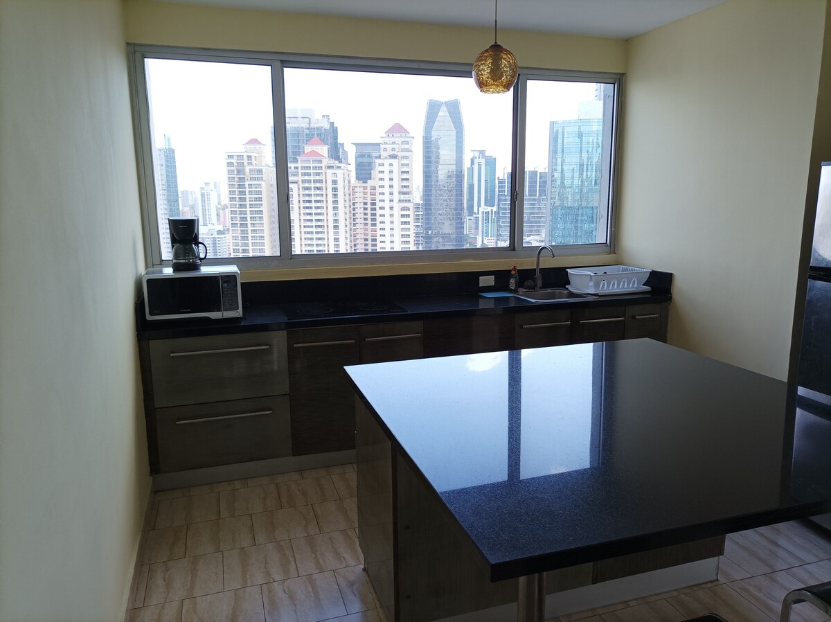 A modern kitchen features sleek cabinets and a spacious island at its center. Large windows provide a view of the city skyline, enhancing natural light in the area. Essential appliances, including a microwave and coffee maker, are visible on the countertop.