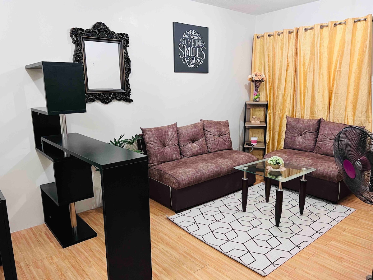 A comfortable seating area is featured with a dark sectional sofa and a glass coffee table, arranged on a patterned rug. A decorative mirror and a shelf with plants are visible, while golden curtains frame the window, allowing light to brighten the space.
