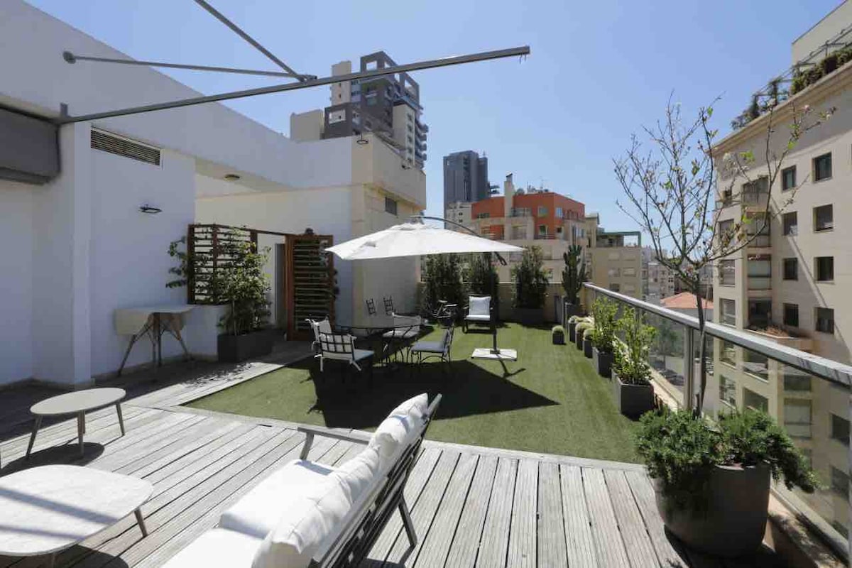 A spacious rooftop terrace offers a green lawn surrounded by potted plants and comfortable seating arrangements. A large umbrella provides shade over a dining area, while lounge chairs invite relaxation under clear blue skies. The cityscape serves as a backdrop, enhancing the outdoor setting.