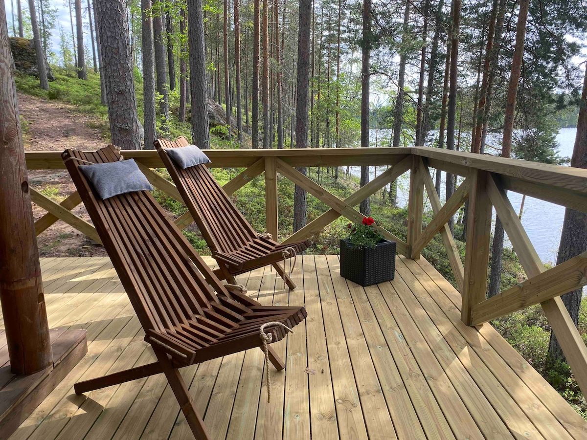 Two wooden lounge chairs are positioned on a deck surrounded by tall trees. A potted plant with red flowers adds a touch of color. The tranquil lake is visible beyond the railing, enhancing the serene setting.