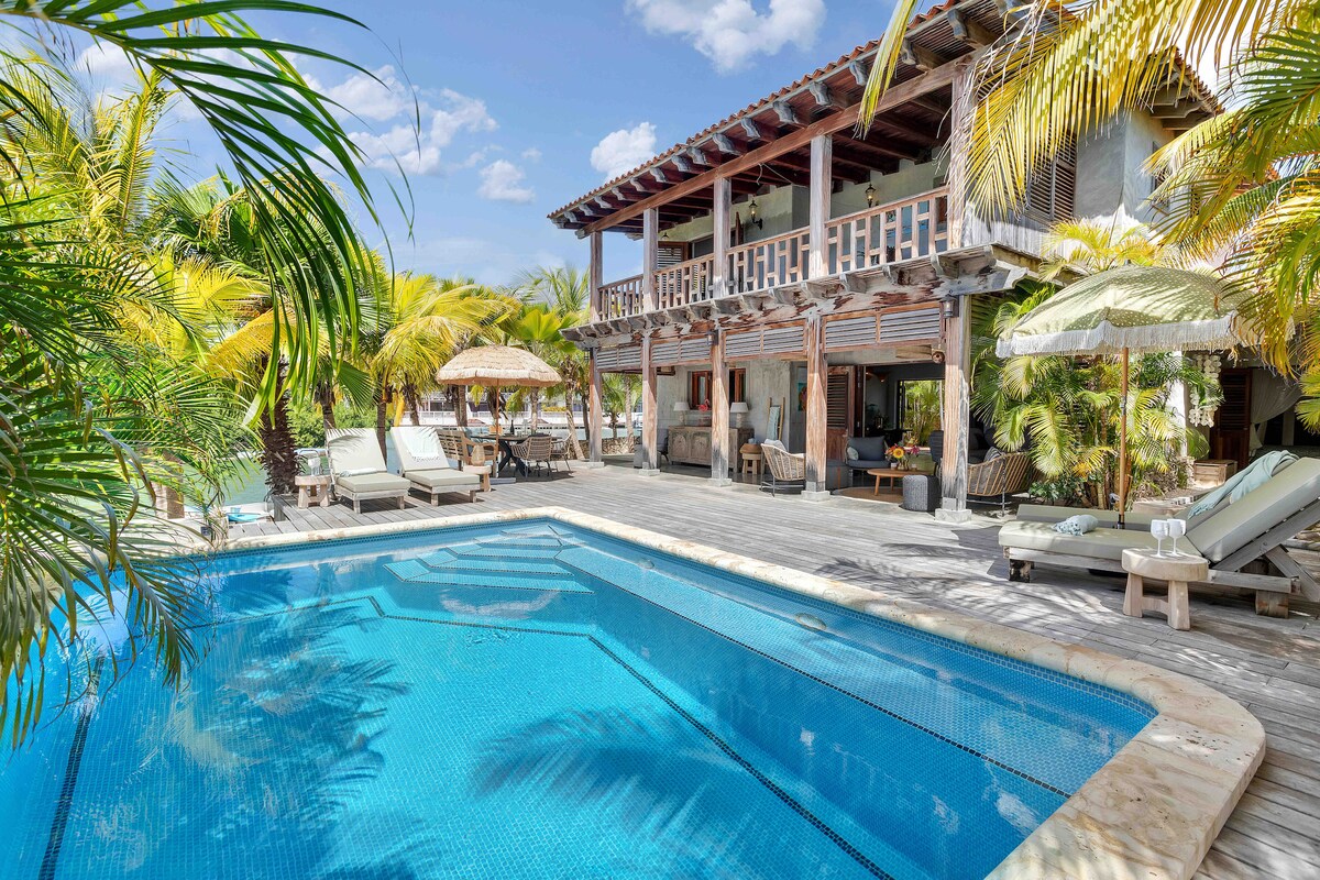 A tropical villa is showcased with a large swimming pool at its forefront, surrounded by lush palm trees. Sun loungers are positioned around the pool, along with shaded seating areas under umbrellas. The structure features a second level with a wide balcony overlooking the outdoor space.