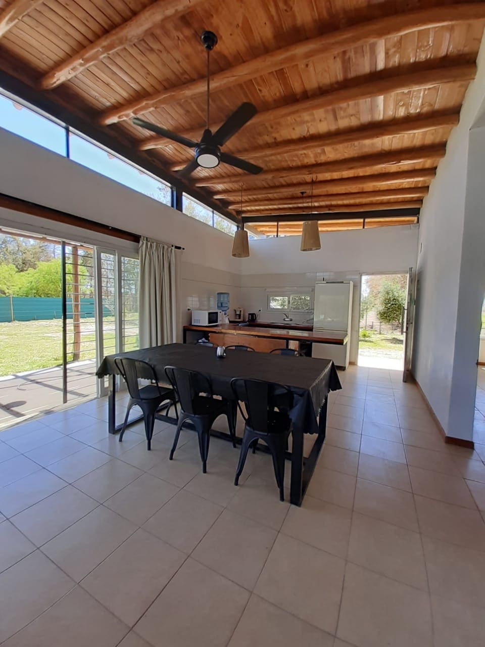 A spacious dining area is illuminated by natural light filtering through large glass doors. A dark table surrounded by several chairs is central to the room, complemented by a ceiling fan and wooden beams overhead. The kitchen area features modern appliances in the background.