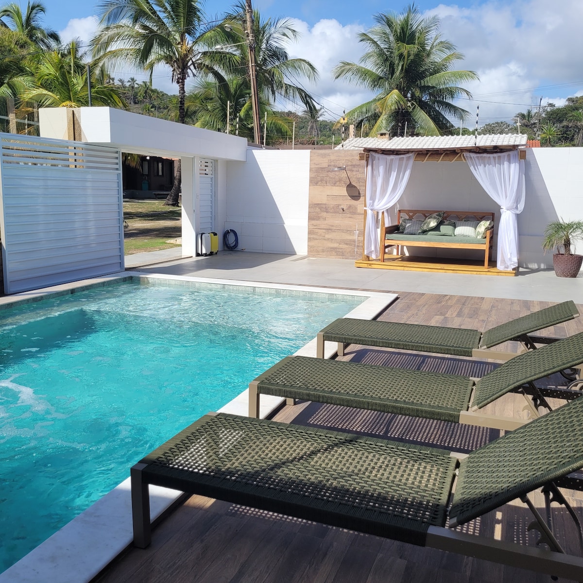 A refreshing pool is surrounded by lounge chairs positioned for relaxation. Beside the pool, a shaded seating area with comfortable cushions invites leisure under the palm trees. The area is enclosed for privacy, enhancing a serene atmosphere.