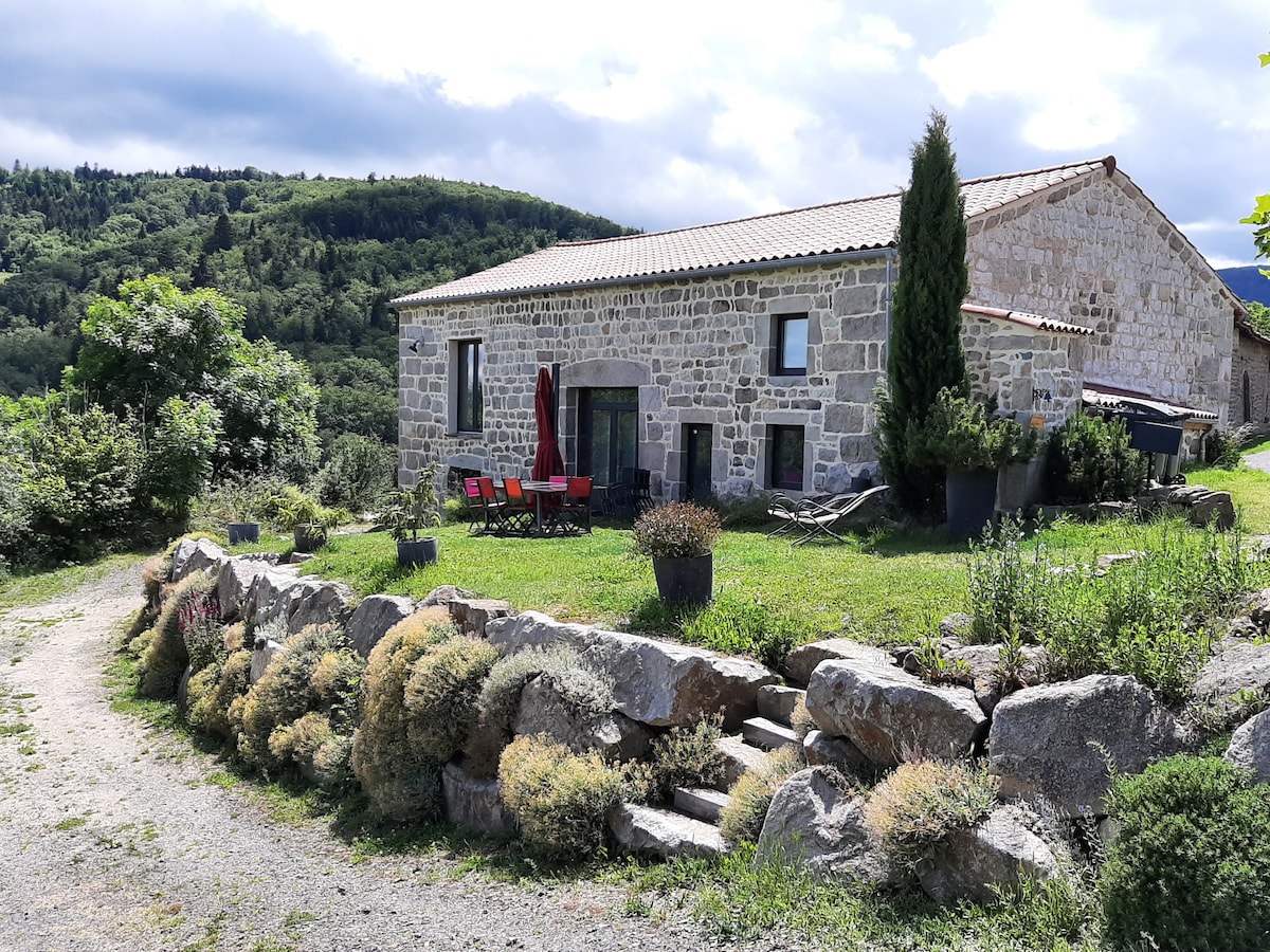 A traditional stone house is set against a backdrop of lush greenery, with a sloped pathway leading to the entrance. A small patio area is visible, featuring a red umbrella and comfortable seating. Decorative plants and low stone walls add texture to the outdoor space.