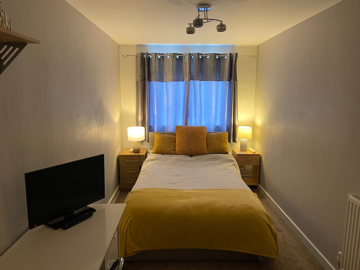 A cozy bedroom is presented, featuring a spacious bed with a yellow duvet and decorative pillows. Two bedside tables with lamps provide soft lighting. A small desk is positioned in the corner, accompanied by a television. Natural light filters through the blue curtains.