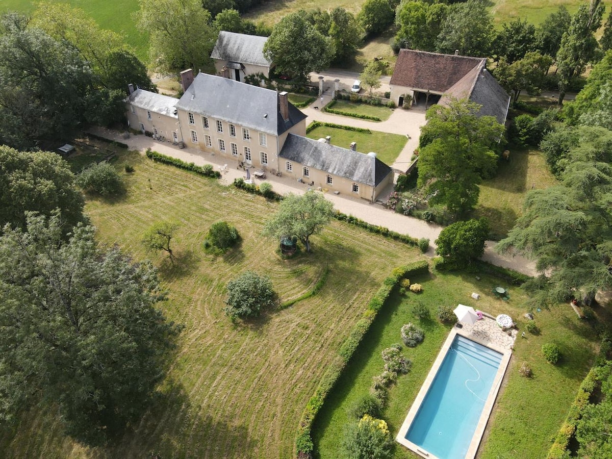 An aerial view showcases a spacious property surrounded by lush greenery and trees, featuring a neatly maintained lawn. A rectangular swimming pool is nestled within the garden, inviting relaxation. Alongside the main building, additional structures and pathways enhance the outdoor scenery.
