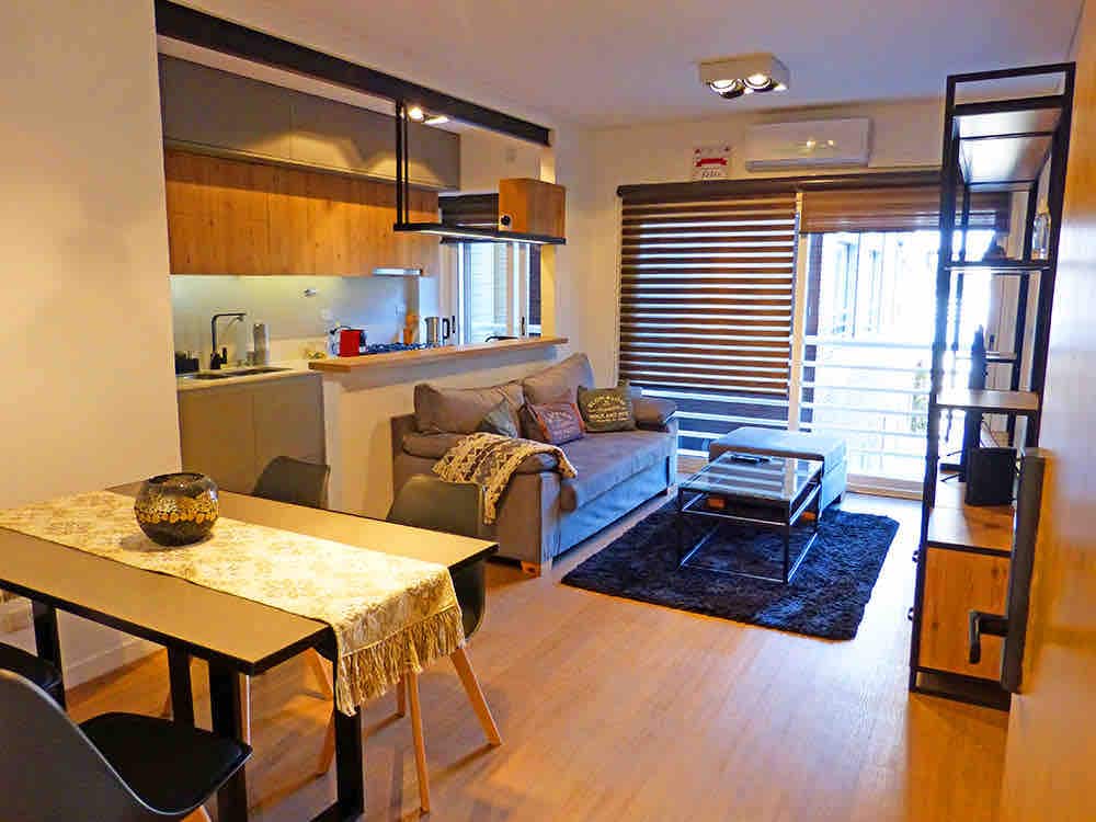 The living area is illuminated by natural light filtering through the window with blinds. A comfortable sofa is paired with a coffee table on a plush rug. A dining table for four is complemented by a decorative centerpiece, and the kitchen space is visible in the background.