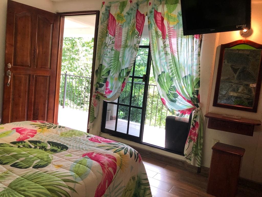 A bright room features large windows adorned with vibrant floral curtains that allow natural light to illuminate the space. A spacious bed is dressed with a colorful patterned cover. A wooden dresser and wall-mounted television complement the tranquil ambiance.