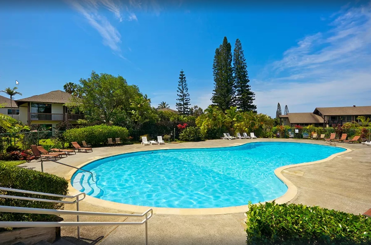 A large swimming pool is surrounded by tropical landscaping, featuring lounge chairs arranged along the water's edge. Lush greenery and tall trees border the area, providing a serene atmosphere. The clear blue sky contrasts with the inviting water, creating a refreshing retreat.