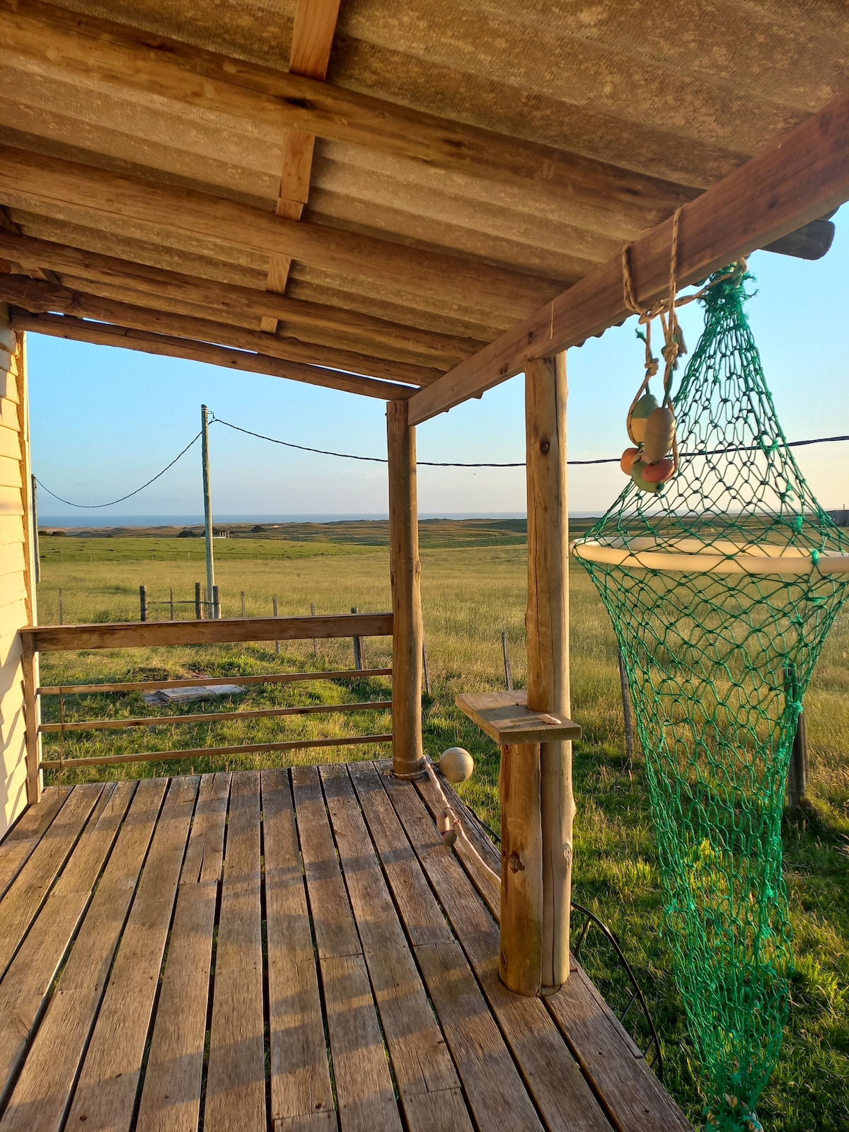 A wooden deck surrounded by natural landscapes offers an unobstructed view of open fields. The sun casts a warm light, enhancing the rustic charm. A green fishing net hangs from the ceiling, adding an authentic touch to the serene setting.