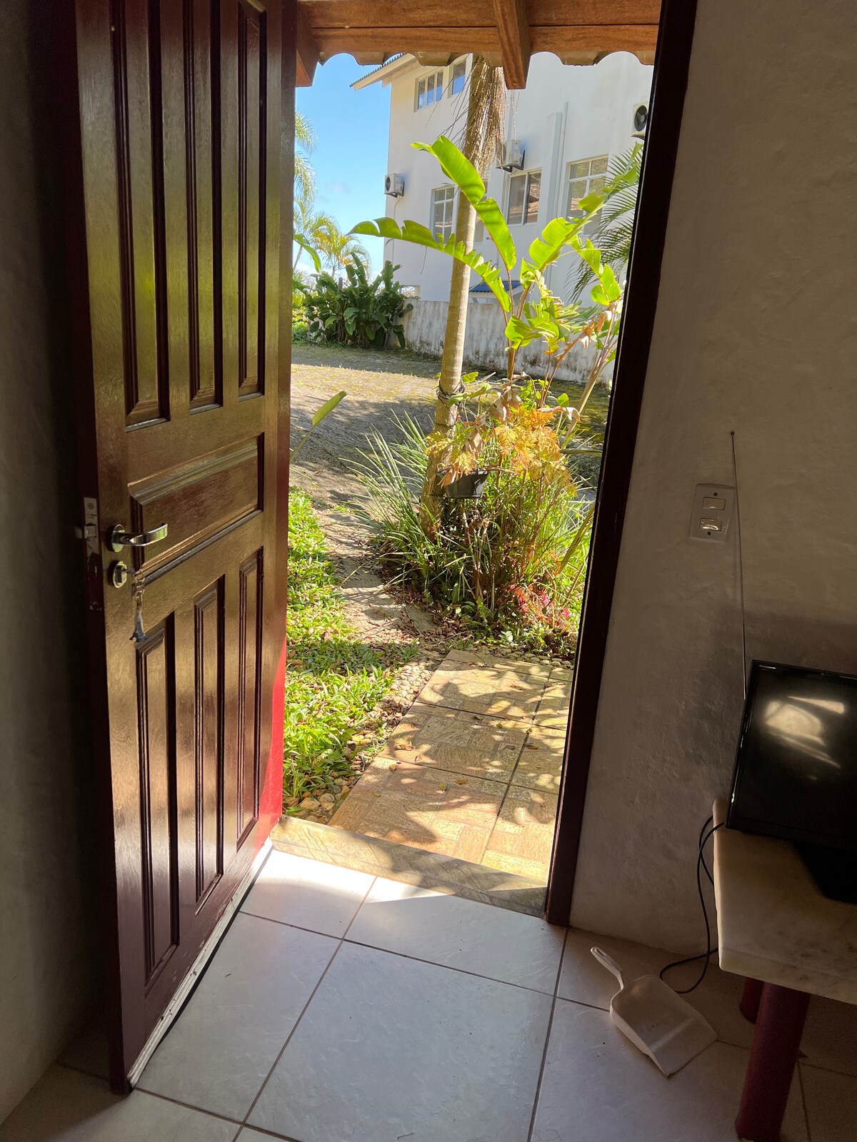 A wooden door stands ajar, revealing a pathway leading to a vibrant garden. Sunlight filters through the greenery, highlighting various plants and stones along the walkway. The interior features a tiled floor and a muted television, providing a glimpse of the outdoor natural setting.
