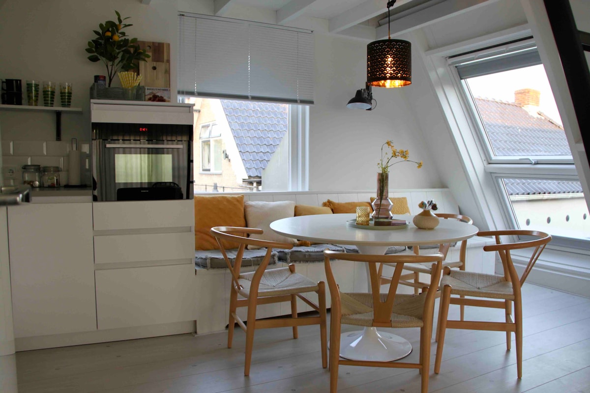 A bright dining area features a round white table with four wooden chairs. A cozy bench with orange cushions lines the wall beneath angled windows. A pendant light casts a warm glow, and a small vase with flowers adds a touch of nature to the setting.