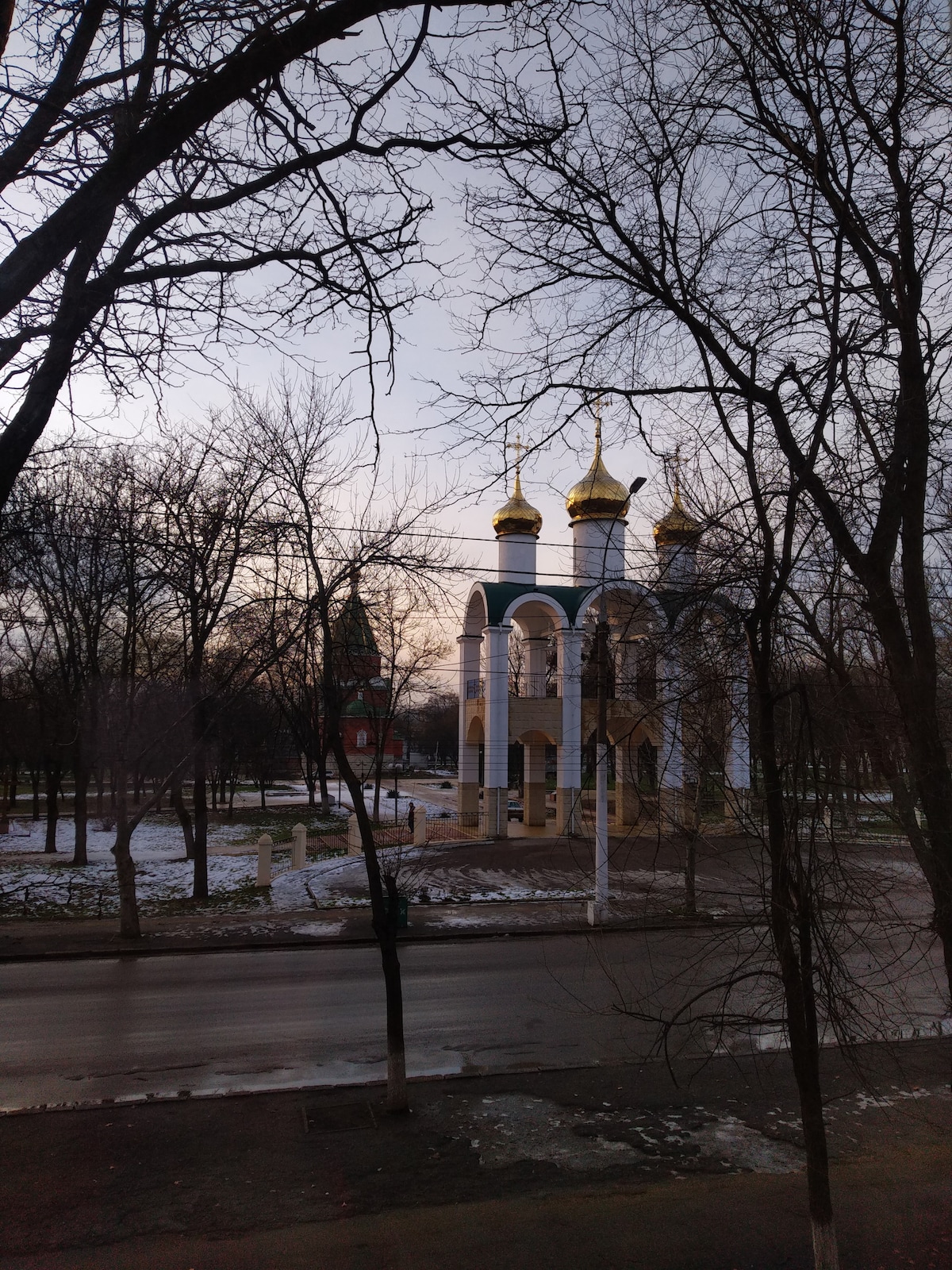 The image captures a view of a church building featuring three golden domes, framed by bare trees. The structure is surrounded by a quiet street and a few patches of lingering snow on the ground, suggesting early spring. Soft sky colors hint at dawn or dusk.