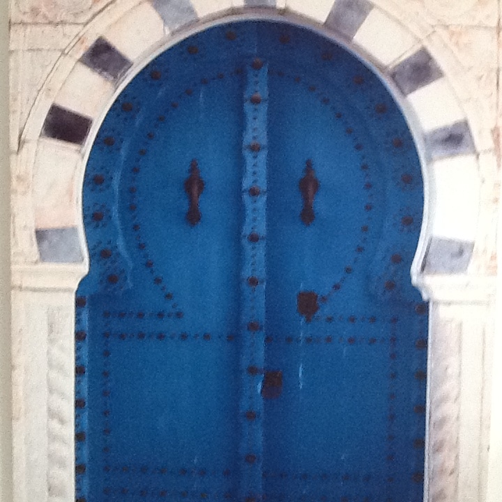 A striking blue door features intricate detailing and a rounded arch design. The surface is adorned with decorative studs, and dual handles provide an inviting entrance. The stone wall surrounding the door offers a contrasting texture, adding to the door's character.