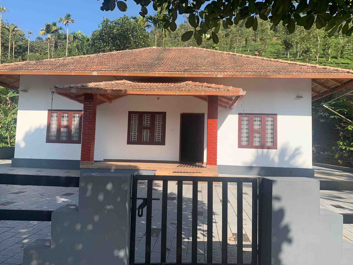 A traditional villa is showcased, featuring a tiled roof and a welcoming entrance flanked by symmetrical windows. The surrounding landscape includes tea plantations, creating a serene environment. A stone pathway leads to the main entrance, enhancing accessibility.