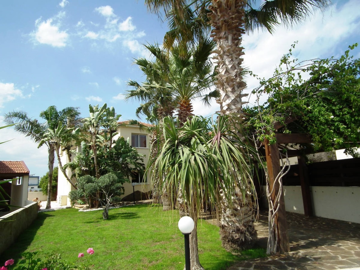 A landscaped garden is featured, showcasing various palm trees and lush greenery. A pathway made of stone leads through the yard, where a round white light fixture stands. The villa, partly visible in the background, is surrounded by a tranquil, natural setting.