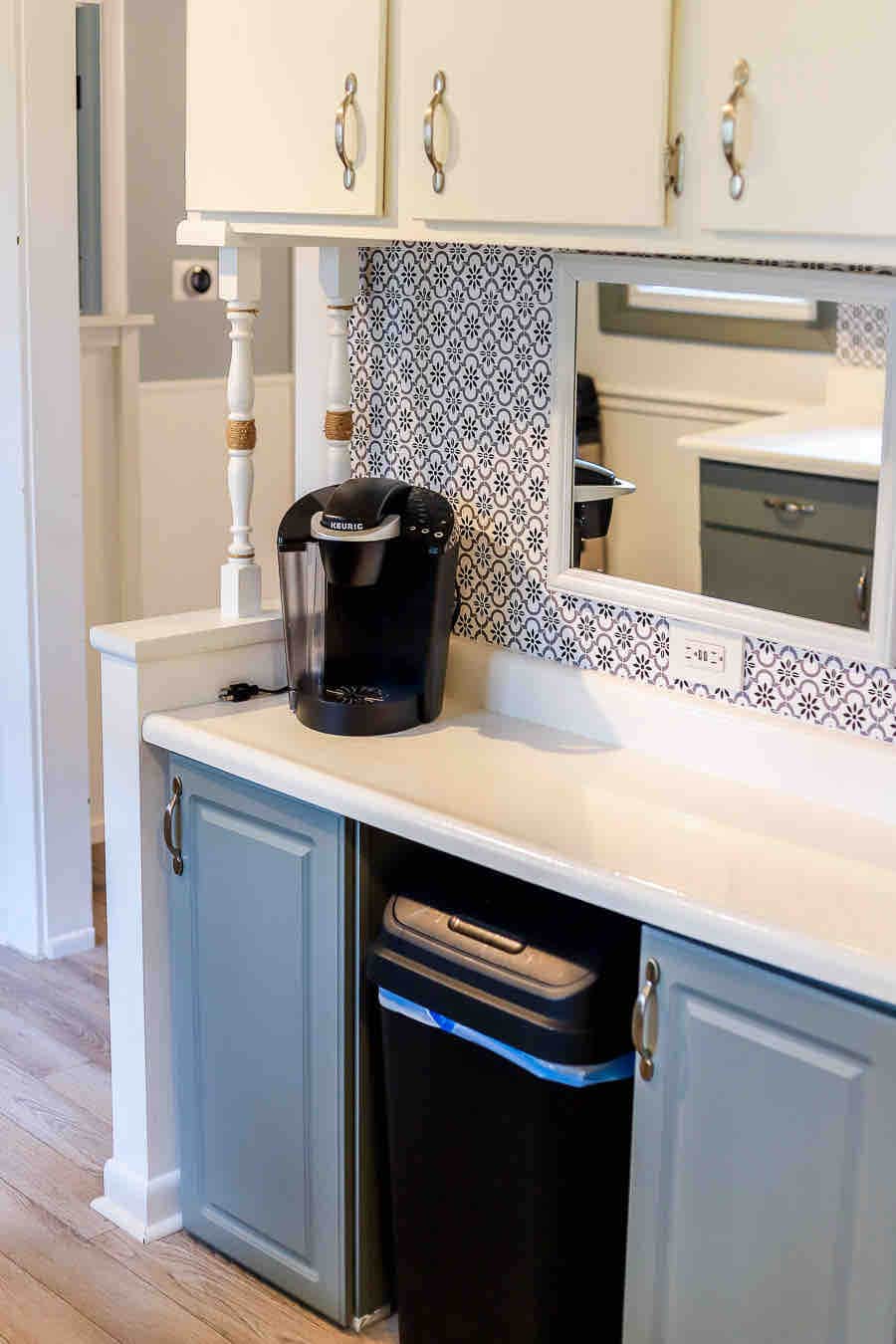 A modern kitchen area features a coffee maker positioned on a counter next to a waste bin. Light-colored cabinetry with decorative handles accents the space, along with a patterned backsplash that adds visual interest. A mirror above the counter reflects the kitchen layout.