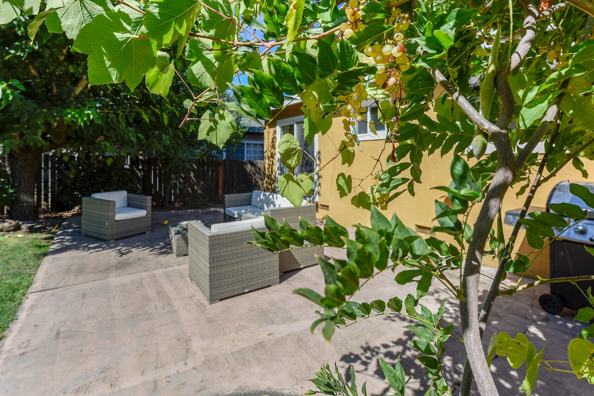 A private backyard patio is bordered by lush greenery. Comfortable seating arrangements are provided with two light-colored chairs positioned around a circular table. A barbecue grill is conveniently placed nearby, enhancing the outdoor dining experience.