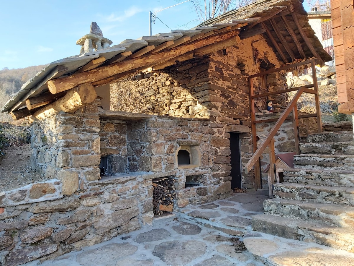 An outdoor cooking area is constructed from rustic stone, featuring a built-in pizza oven and a spacious cooking area. A wooden structure with a sloped roof is complemented by a stone pathway leading to the entrance. Natural surroundings enhance the peaceful setting.