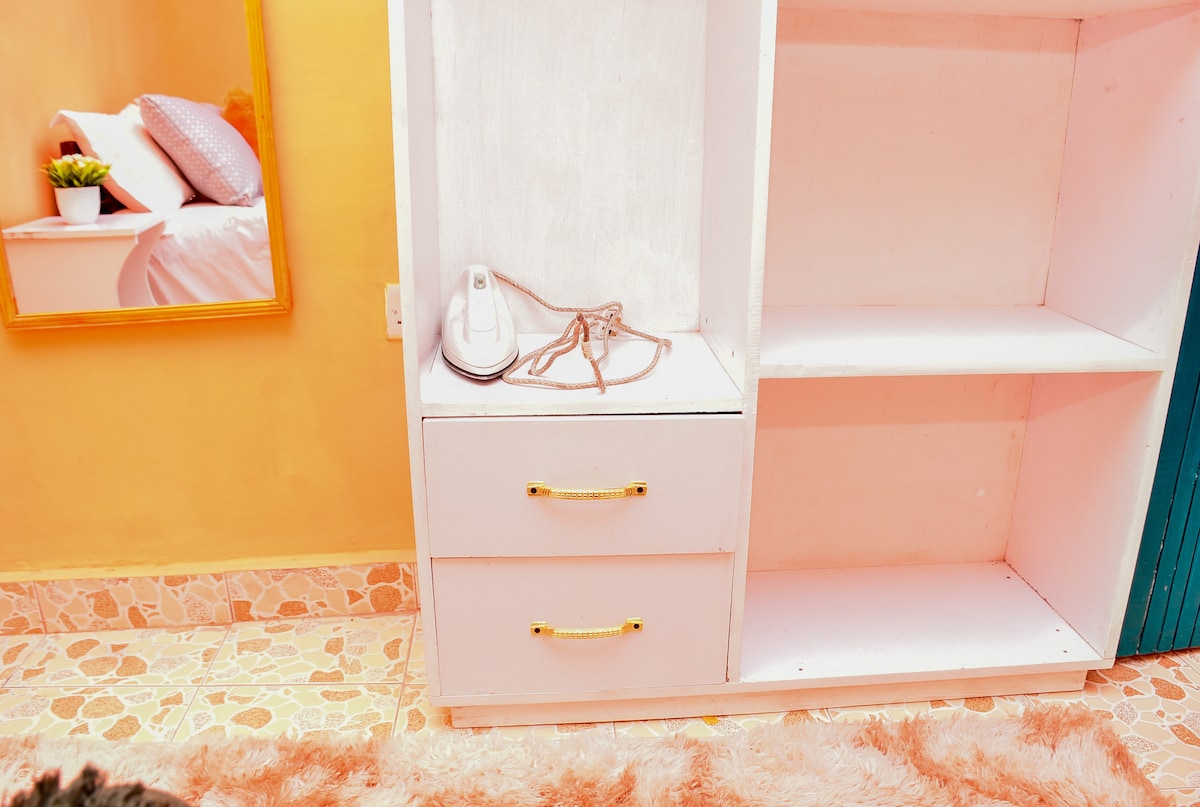 A functional storage area is presented, featuring a white cabinet with two drawers. An iron, accompanied by a cord, rests on the surface. The backdrop displays a mirror, and a texture-rich rug complements the area, enhancing the inviting ambiance.