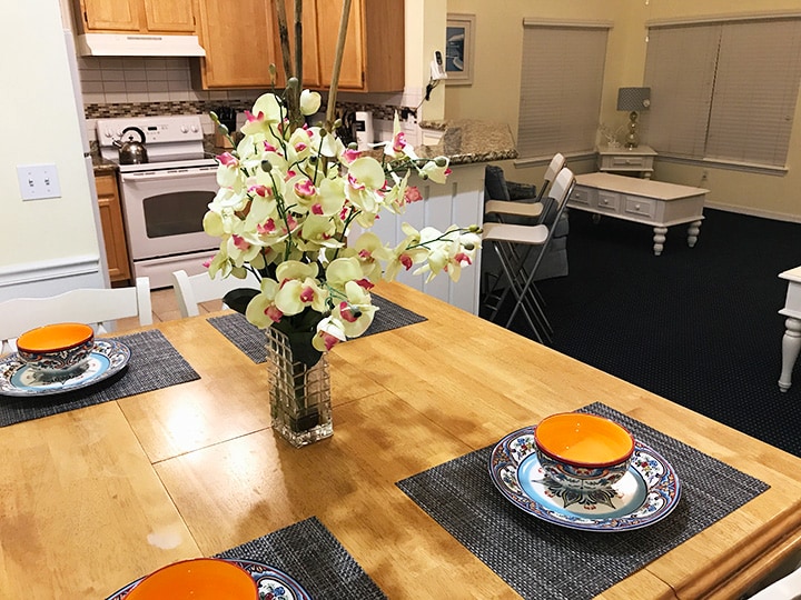 A dining table is set with contrasting orange bowls and decorative plates, surrounded by an arrangement of artificial flowers in a glass vase. The kitchen area with wooden cabinetry and appliances is visible in the background, along with a spacious living room that features comfortable seating.