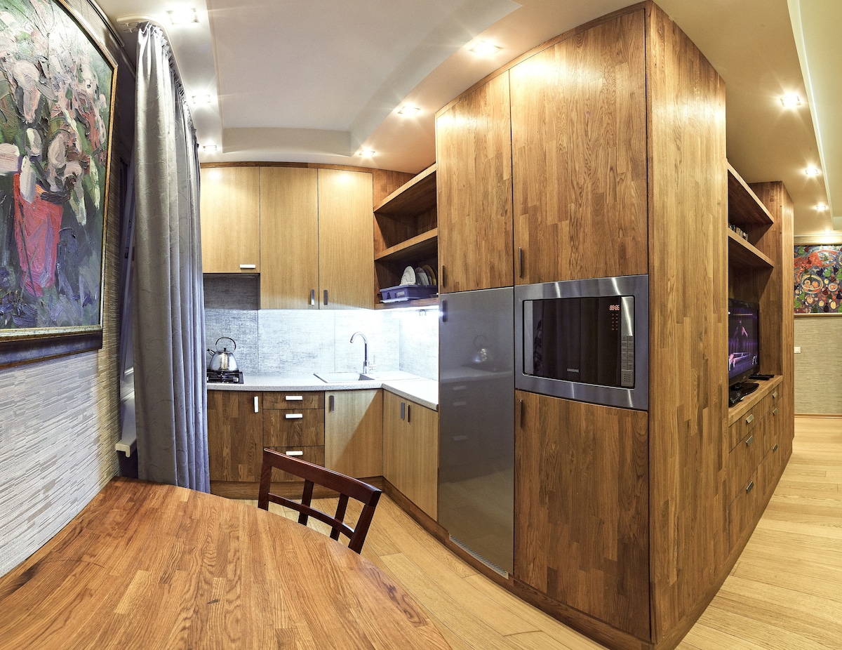 A kitchen area is characterized by warm wood cabinetry and sleek countertops. The layout includes built-in appliances, including a microwave and a refrigerator. Under-cabinet lighting highlights the workspace, while a wooden dining table with a chair provides a casual dining option.