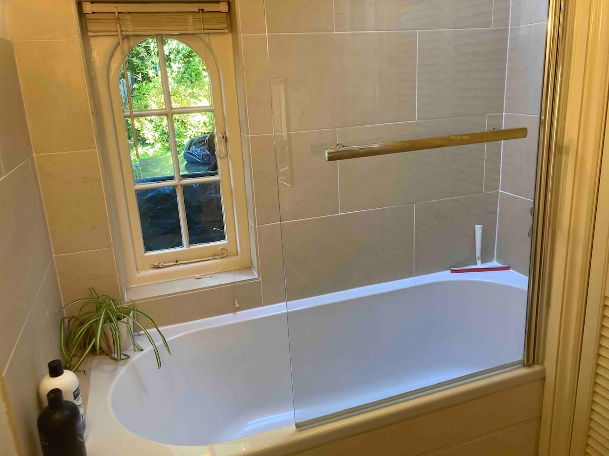 An ensuite bathroom features a large bathtub with a glass shower screen. Natural light enters through an arched window, highlighting the neutral tiled walls. A small plant sits on the ledge, and various toiletries are neatly arranged nearby.