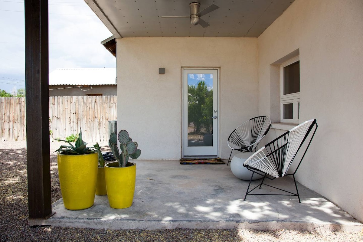 An outdoor porch area features two decorative chairs alongside large potted plants. A vibrant yellow pot holds a green cactus, and a white pot is filled with foliage. The entrance door is positioned at the center, framed by a simple concrete floor beneath a sheltered roof.