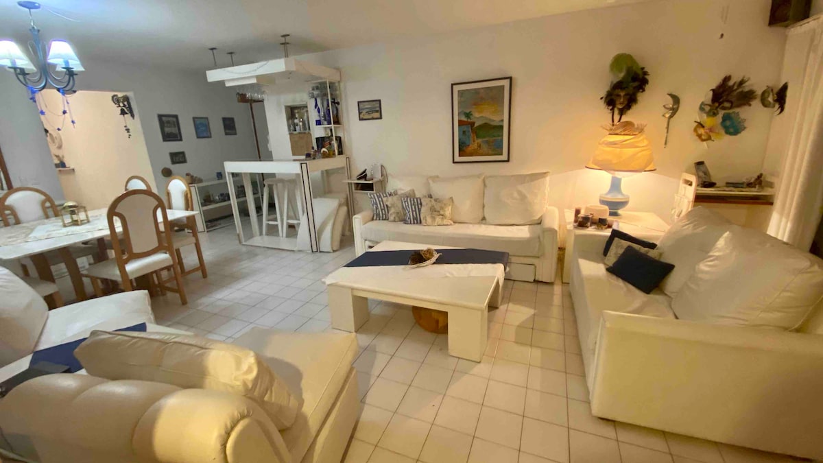 A spacious living area features comfortable, white seating arranged around a central coffee table. A dining area with wooden chairs and a round table is visible in the background. Soft lighting is provided by a blue chandelier, and decorative art hangs on the walls.