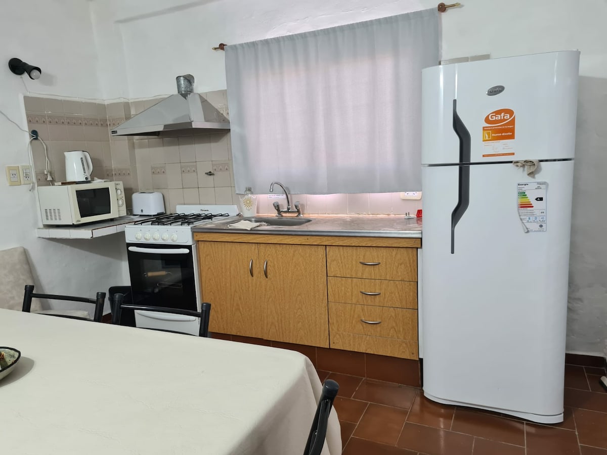 A functional kitchen is presented, featuring a white refrigerator and a sleek gas stove with a range hood above. A microwave sits next to the sink, with wooden cabinetry below. Natural light filters through a curtain-covered window, illuminating a dining table set for gathering.