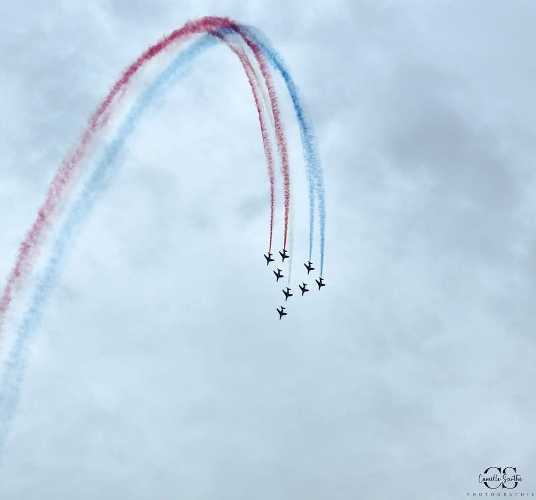 A formation of eight aircraft performs an aerial display, creating trails of red and blue smoke against a cloudy sky. The aircraft are positioned in a dynamic arrangement, showcasing their flight capabilities.