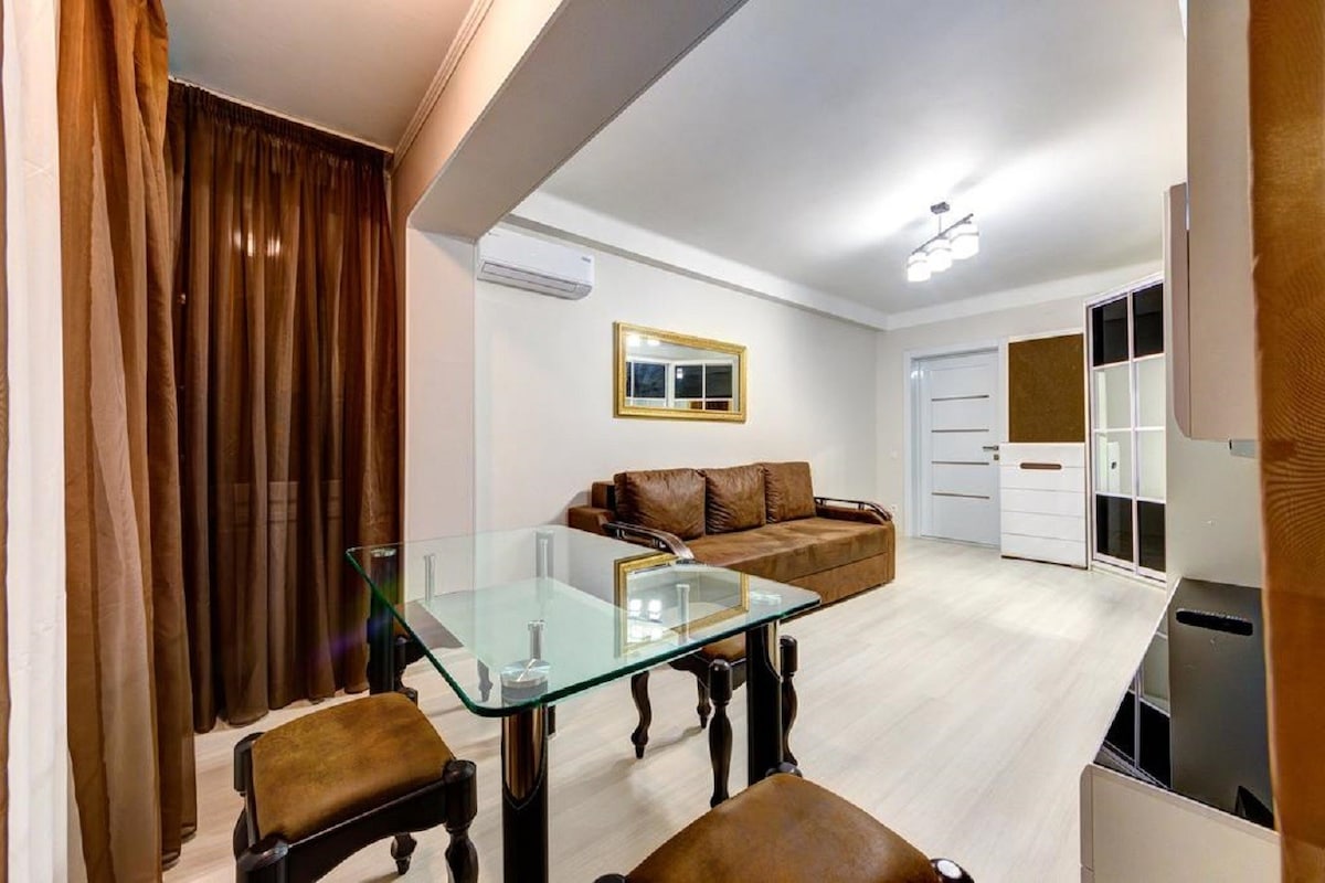 A modern living space features a glass table and brown chairs, complemented by a comfortable brown sofa. Neutral-colored walls are paired with rich brown curtains, while a decorative mirror enhances the room's light. A door leads to another area, providing access to the apartment's layout.