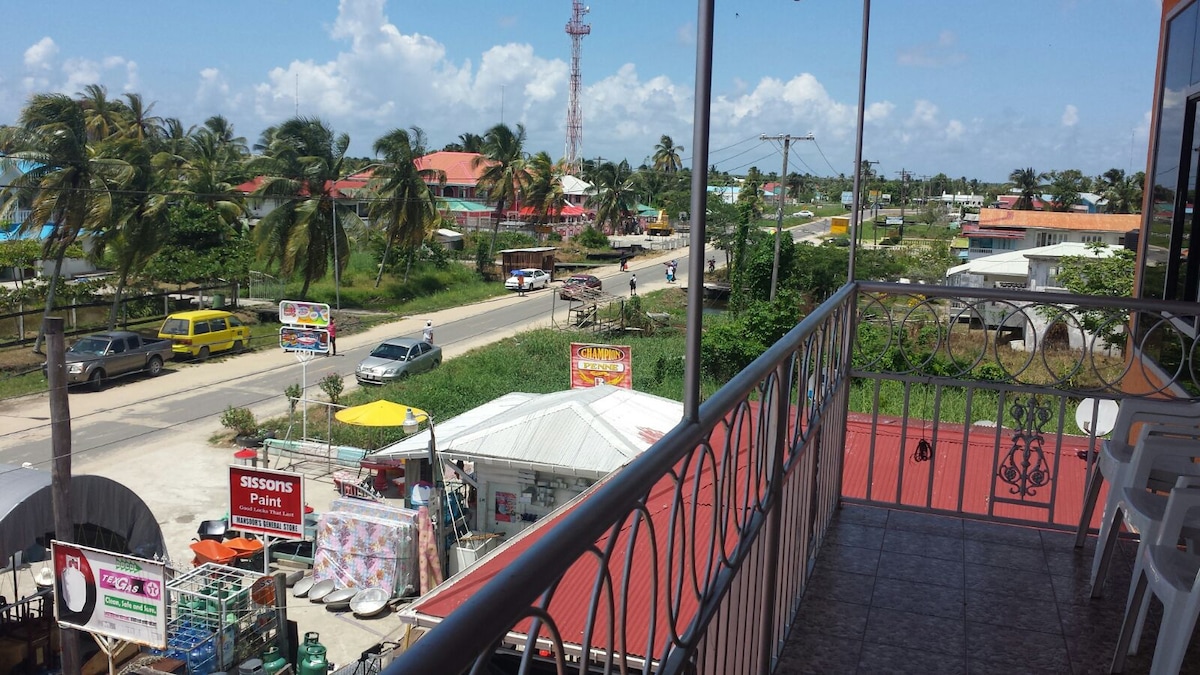 A view from a private balcony overlooks a vibrant street lined with palm trees, colorful buildings, and local shops. A clear sky with scattered clouds provides a bright backdrop to the lively scene below, where people and vehicles create a sense of community.