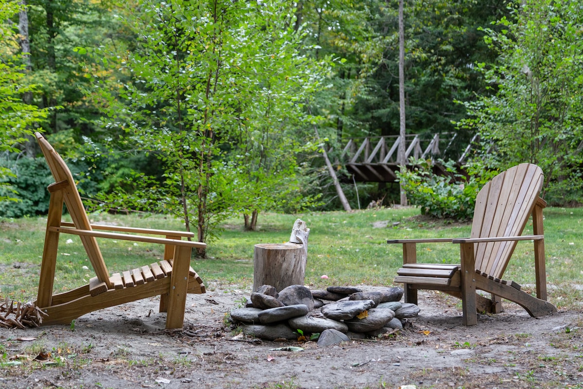 Two wooden adirondack chairs are positioned around a circular arrangement of smooth stones, forming a fire pit. A natural wooden stump rests in the center, surrounded by a grassy area, with trees providing a peaceful backdrop.