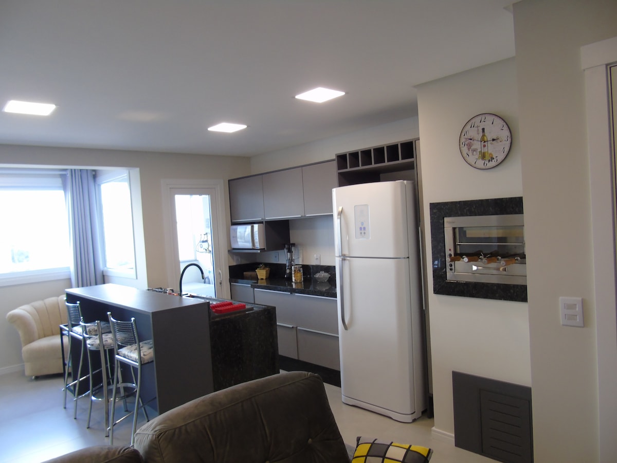 The modern kitchen features a refrigerator and built-in oven, complemented by a sleek countertop with bar seating for four. An open layout connects the kitchen to a cozy living area, illuminated by ample natural light from nearby windows.