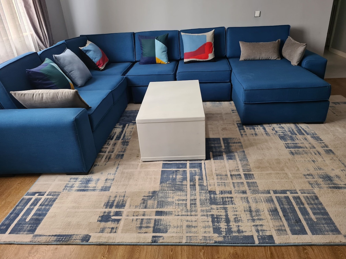 A spacious blue sectional sofa is arranged comfortably in a living area, complemented by colorful throw pillows. A light-colored coffee table sits in the center, resting on a patterned area rug featuring shades of beige and blue.