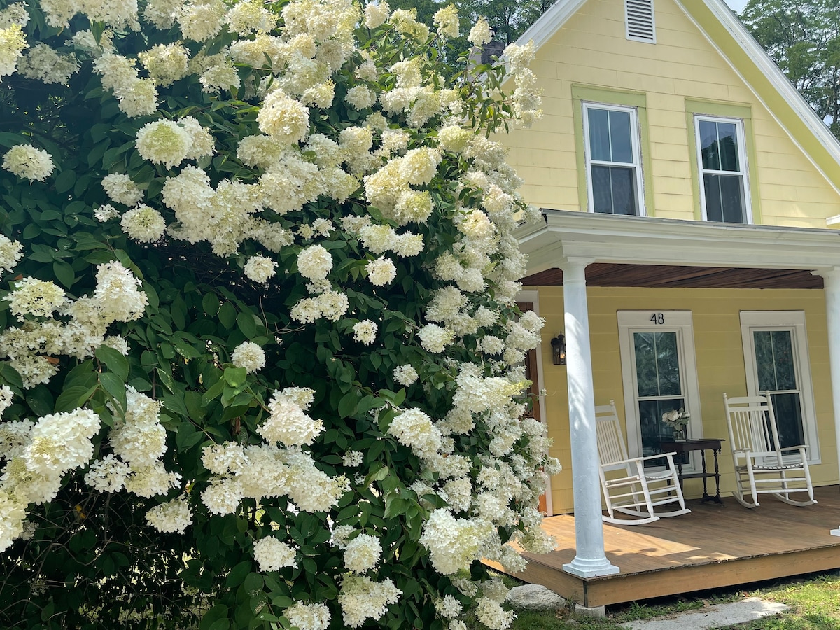 A charming yellow house features a welcoming front porch with two rocking chairs. Lush white hydrangea blooms cascade from a nearby bush, enhancing the inviting atmosphere of the entryway.