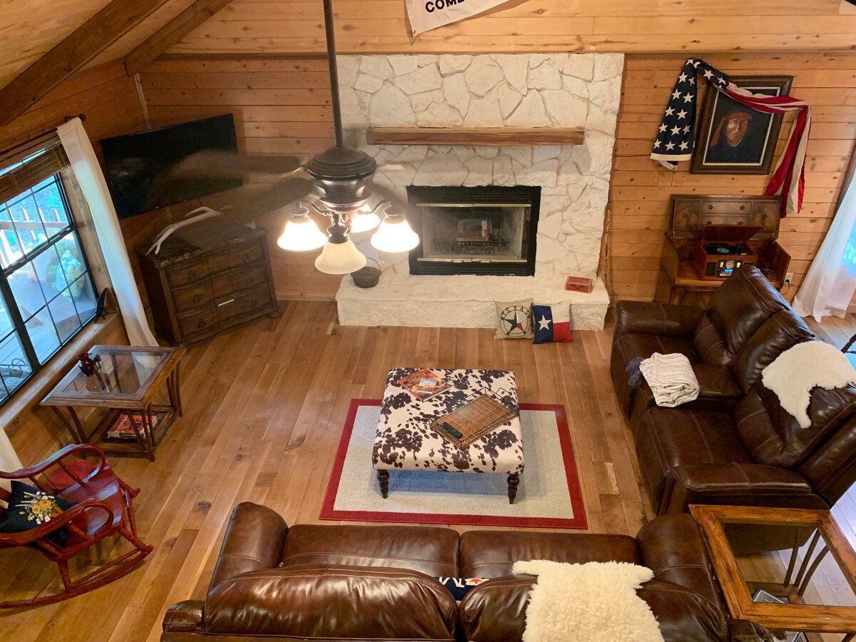 The living room features a spacious seating arrangement with dark leather sofas and a coffee table at the center. A stone fireplace is set against the wooden wall, while large windows fill the space with natural light. Rustic decor and wooden accents create a comfortable ambiance.
