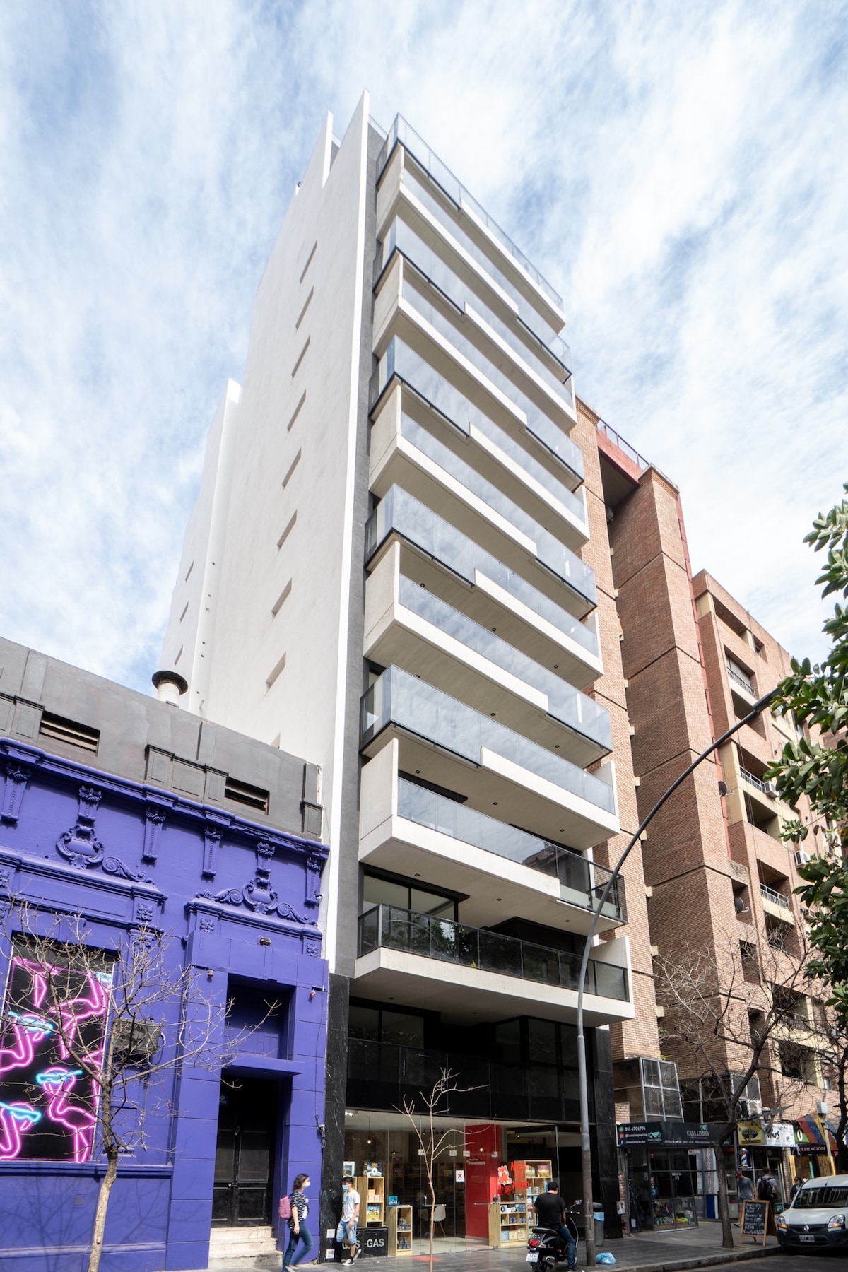 The modern building showcases a sleek architectural design with five levels and expansive balconies. Glass railings provide unobstructed views, while the white exterior contrasts with the colorful adjacent structure. The ground floor features large windows, inviting natural light into the commercial space.