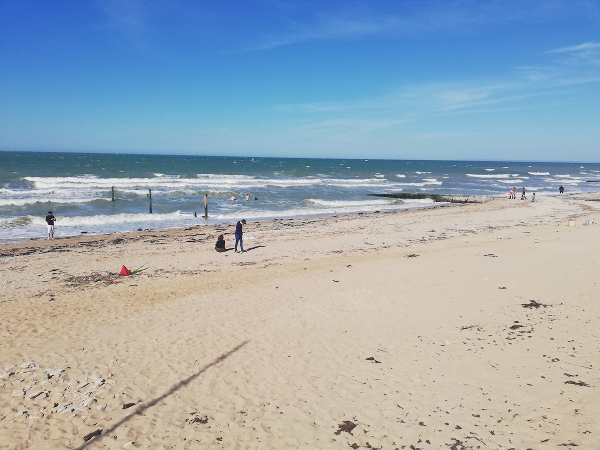 A sandy beach is shown with gentle waves lapping at the shore. Several individuals can be seen walking along the water's edge, while distant wooden posts emerge from the sea. The sky is clear, decorated with a few scattered clouds.