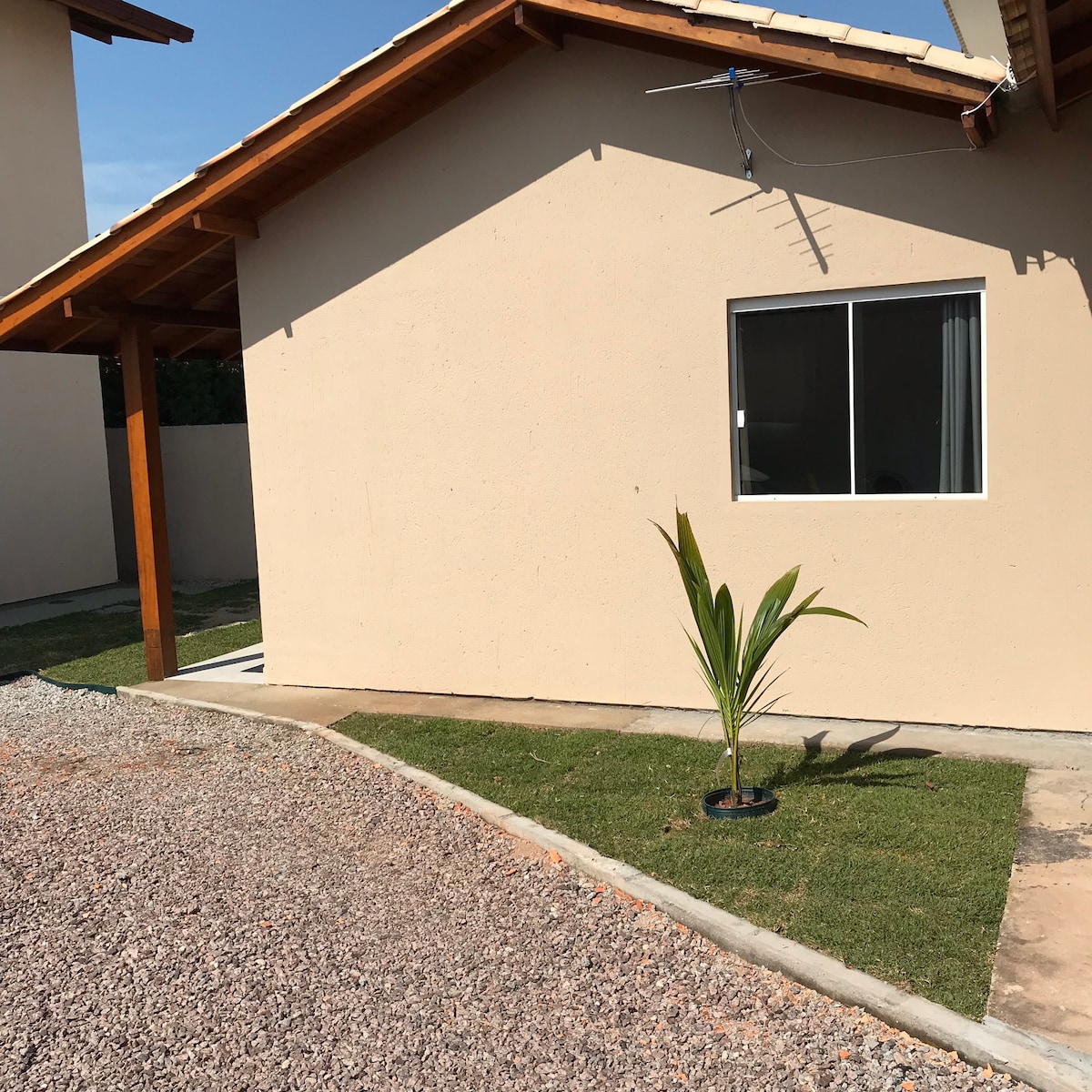 A light-colored exterior of a single-story building is visible, featuring a large window on the front. Green grass is maintained around a small planted palm tree, and a gravel pathway leads up to the entrance, creating a welcoming approach.
