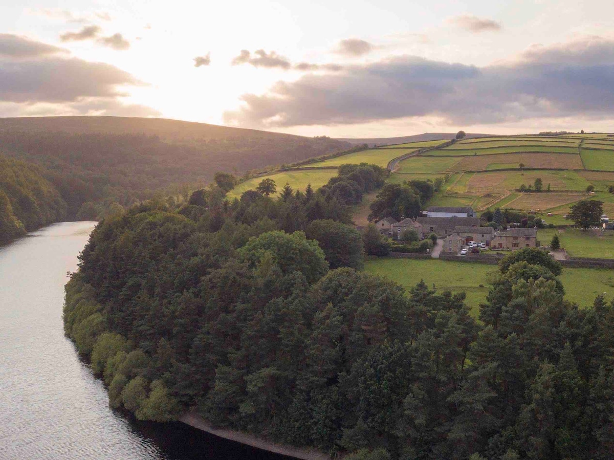 An aerial view captures a serene landscape featuring a winding river bordered by lush greenery and gently sloping hills. The sun sets behind the hills, casting a warm glow over the land and creating a calming atmosphere, with open fields and woodland visible in the distance.
