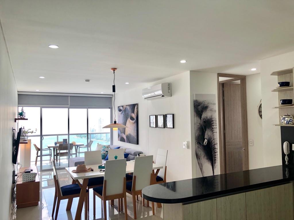 A modern living space features a dining area with a wooden table surrounded by six light-colored chairs. A comfortable seating arrangement is visible in the background, complemented by large windows offering a view of the bay. Soft lighting enhances the neutral decor and artwork.
