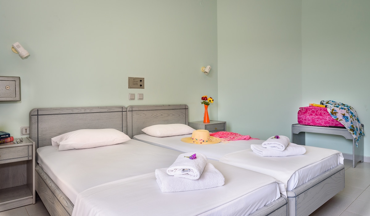 A light and airy bedroom is presented, featuring two spacious beds neatly made with fresh linens. A couple of towels are arranged on the beds. A small nightstand and wall-mounted lights are positioned beside each bed. Decorative items include a hat and colorful textiles displayed on the beds.