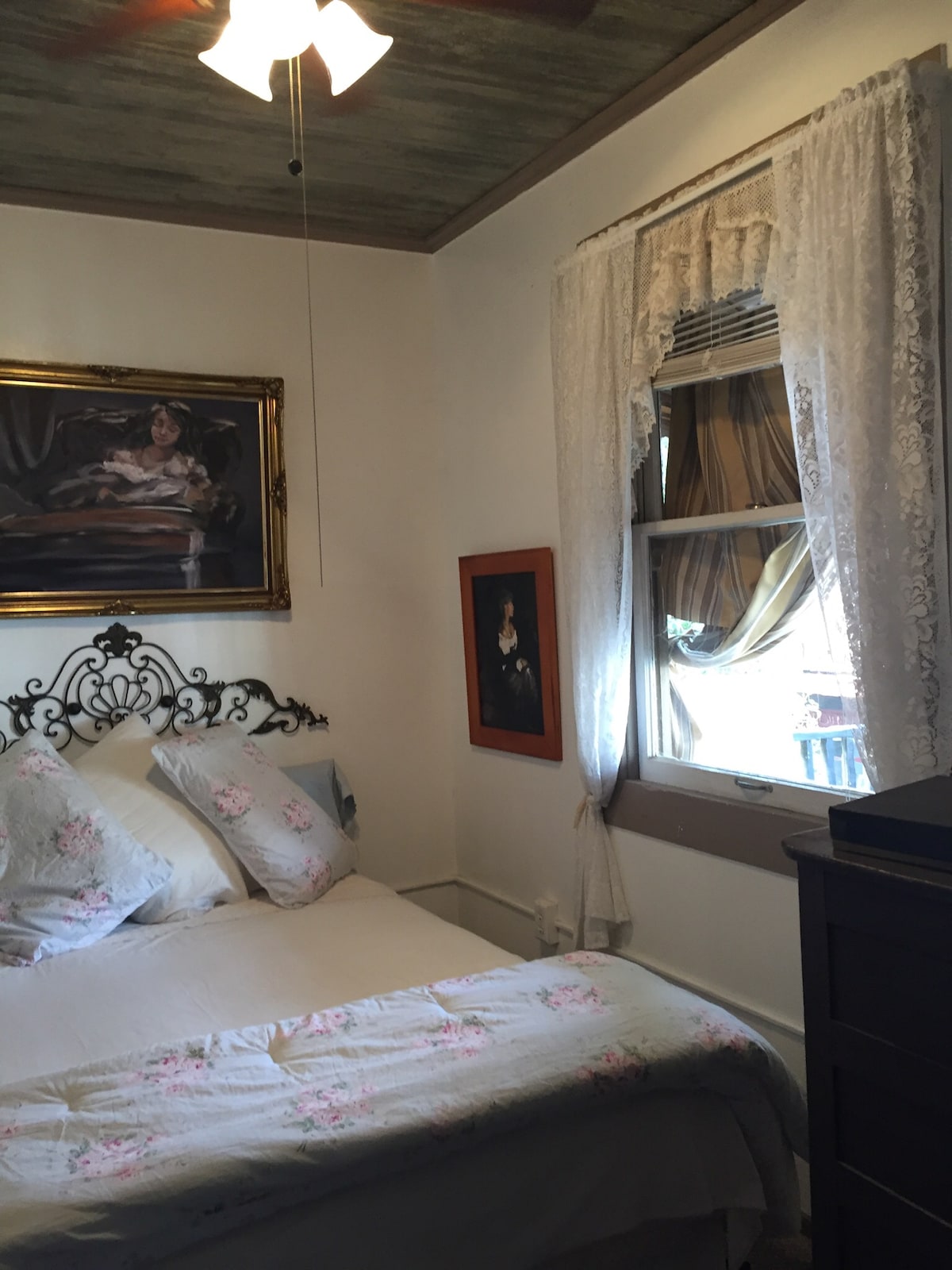 A private bedroom is furnished with a decorative metal bed frame and soft bedding. White lace curtains frame the window, allowing natural light to illuminate the room. A vintage artwork hangs on the wall, complemented by an ornate mirror above the bed.