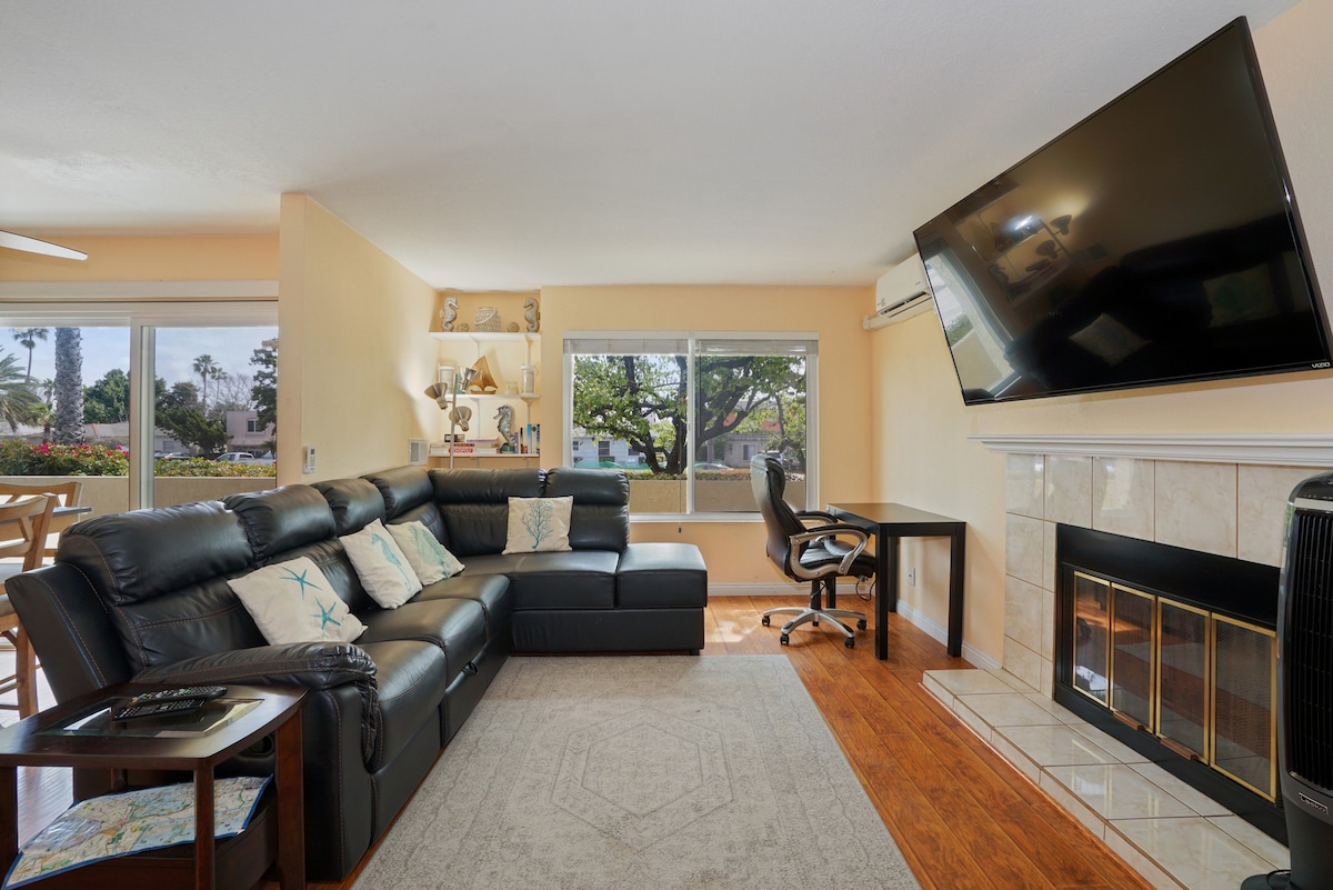 A comfortable living area is presented, featuring a black sectional sofa adorned with decorative pillows. A large flat-screen TV is mounted on the wall above a fireplace. Natural light fills the space through expansive windows, with a desk and chair positioned nearby.