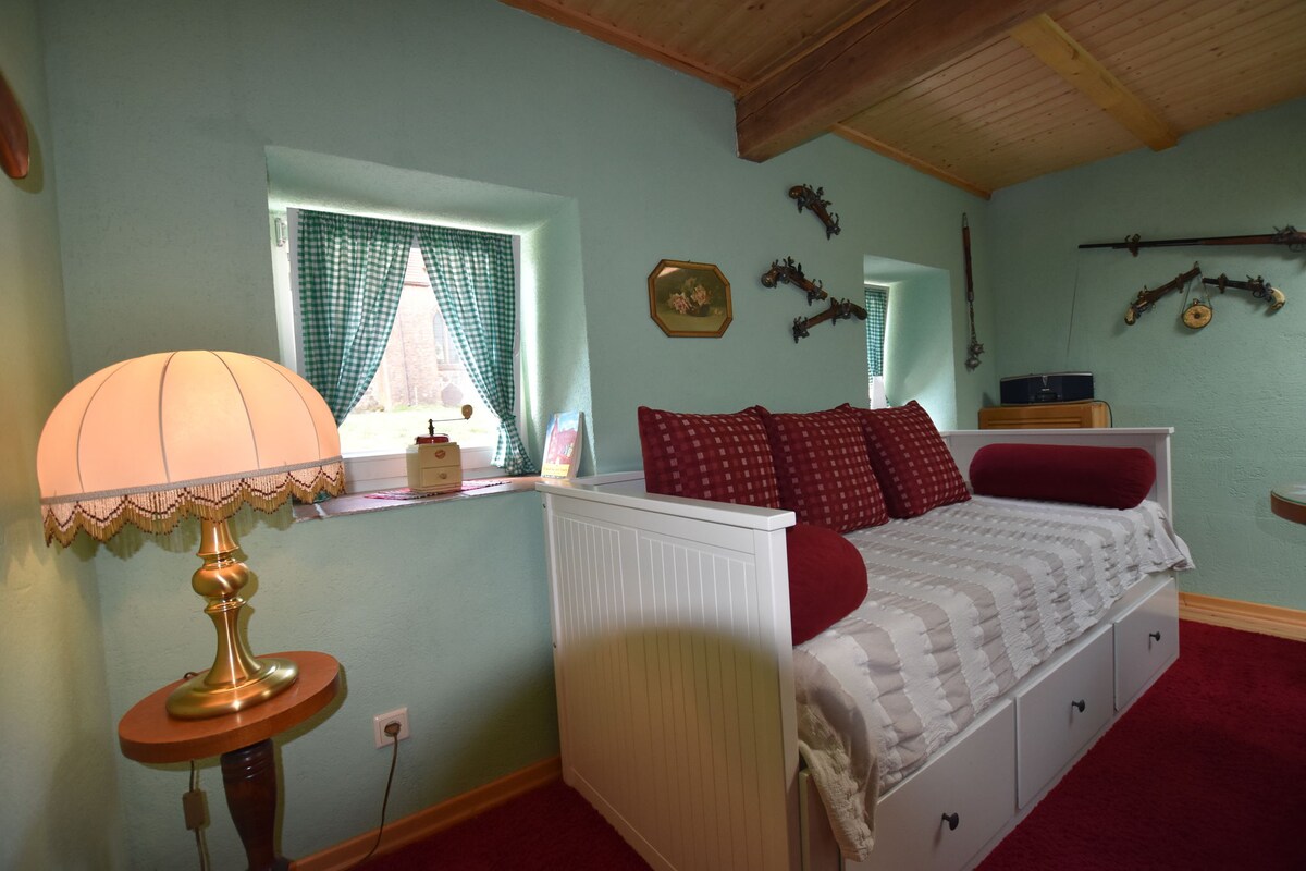 A well-lit area features a cozy daybed adorned with red and white pillows. A lamp with a cream shade stands beside it, providing soft illumination. Two small windows are dressed with green gingham curtains, allowing natural light to enhance the space.