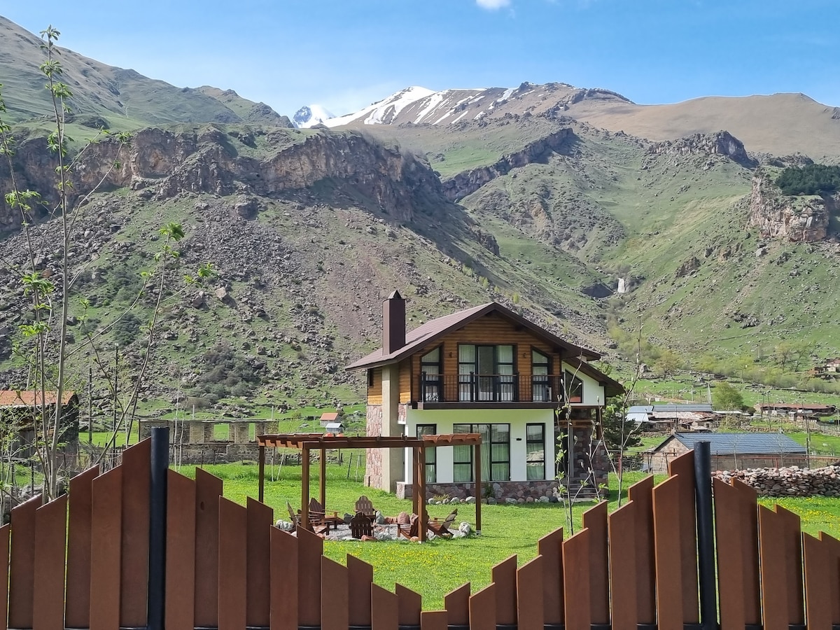 A two-story villa is positioned against a backdrop of towering mountains, some capped with snow. Lush green hills surround the property, while a wooden fence frames the scene. A stone pathway leads to an outdoor seating area, enhancing the tranquil setting.