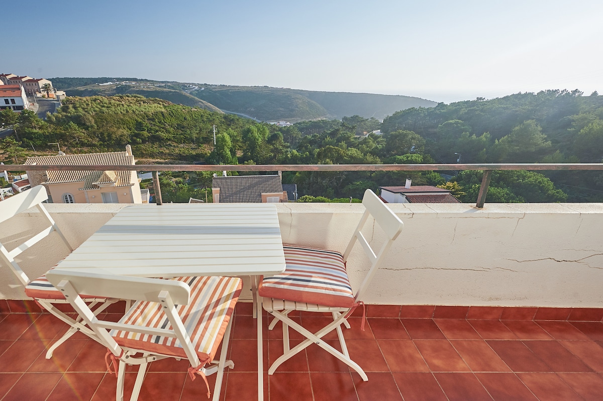 An outdoor balcony is seen, featuring a small white table with two striped chairs. A panoramic view of green hills and a valley stretches into the distance, under a clear blue sky, offering a sense of tranquility.
