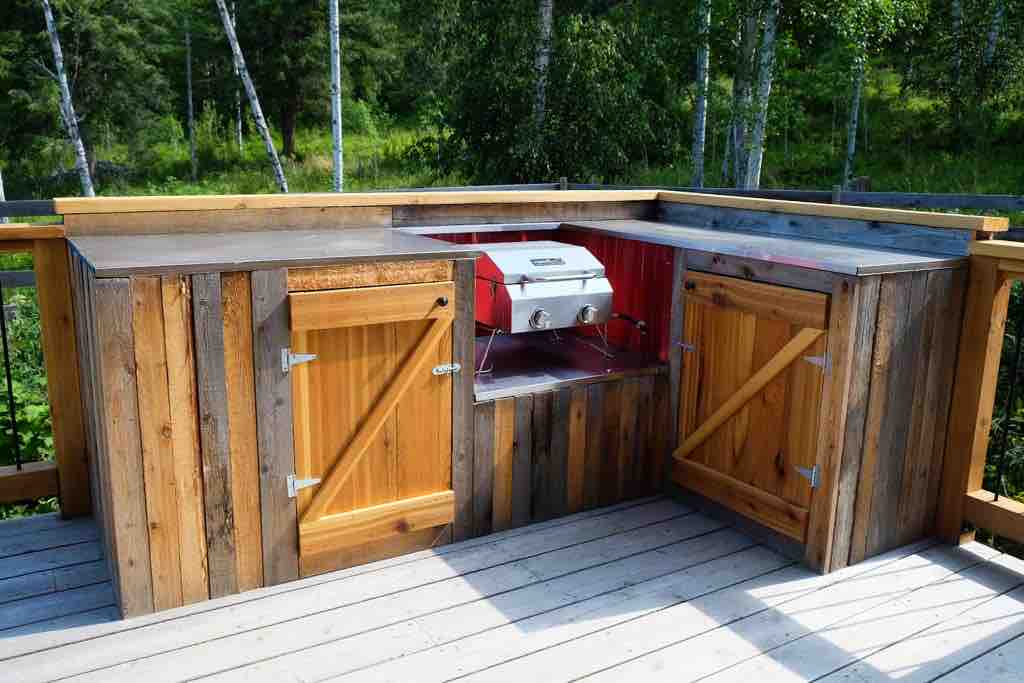 An outdoor grilling area is presented with a sturdy wooden structure featuring a built-in gas grill. Two cabinets with wooden doors provide storage, while a spacious countertop allows for food preparation. Green foliage in the background adds a natural touch to the setting.